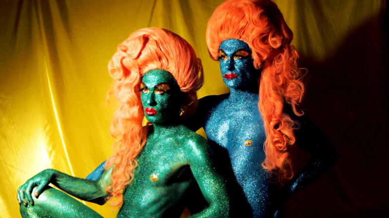 Zwei Personen mit glitzernder blauer und grüner Körperbemalung und orangen, voluminösen Perücken vor gelbem Hintergrund.