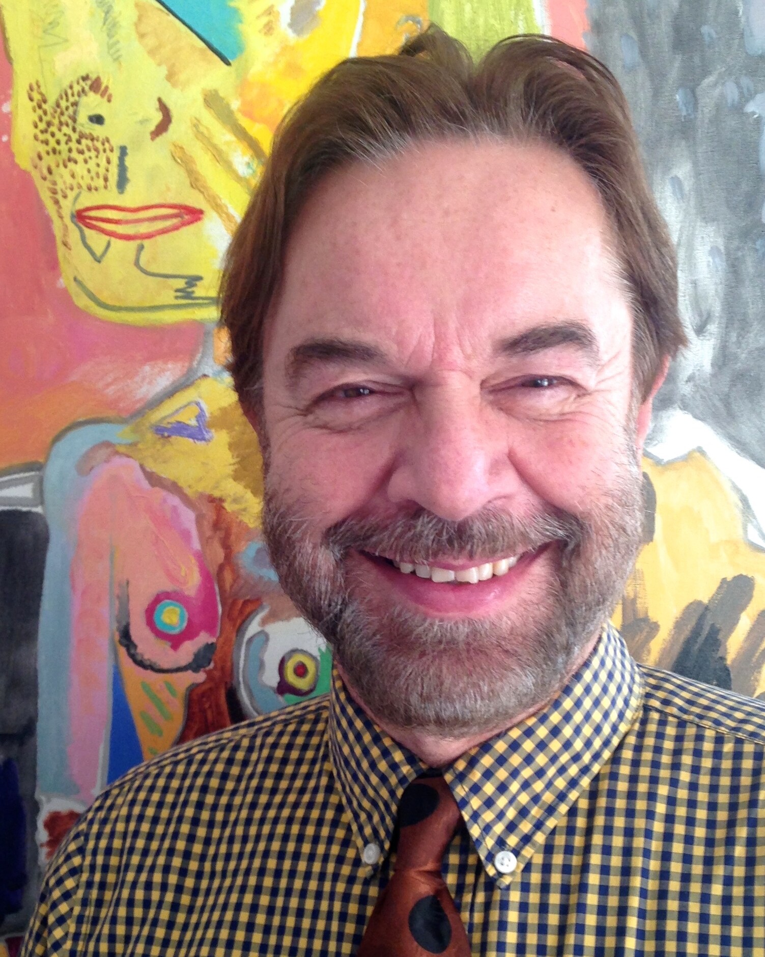 Smiling man with beard wearing checked shirt and polka dot tie, standing in front of colourful abstract painting.