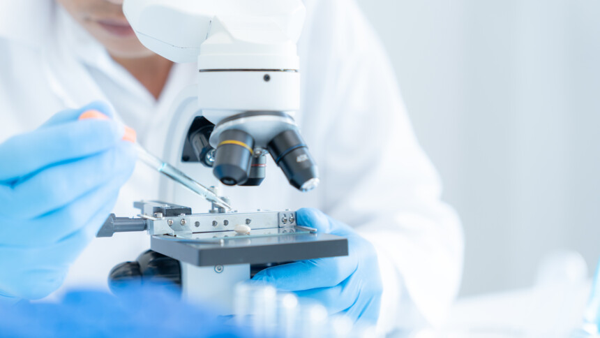 Wissenschaftlerin mit blauen Handschuhen untersucht Probe unter Mikroskop im Labor.