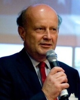 Älterer Mann mit Glatze und roter Krawatte spricht in ein Mikrofon.