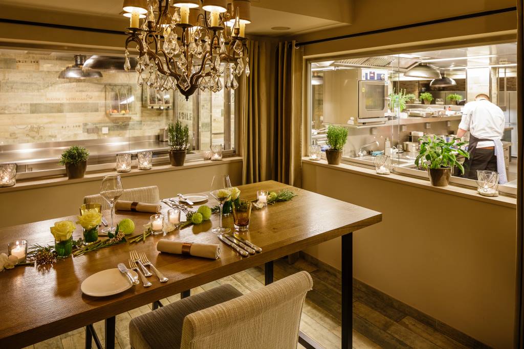 Eleganter Esstisch mit Kerzen und Blumen vor einer Glastür zur offenen Restaurantküche mit Koch.