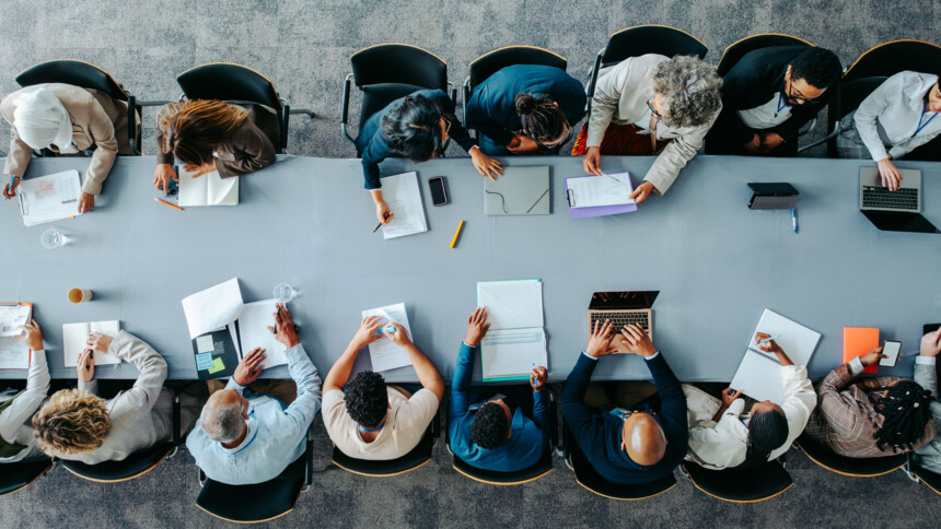 Overhead view of diverse professionals collaborating around a large conference table with laptops and documents.