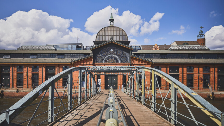 View of a metal pedestrian bridge leading to a historic red brick building with large windows and a dome.