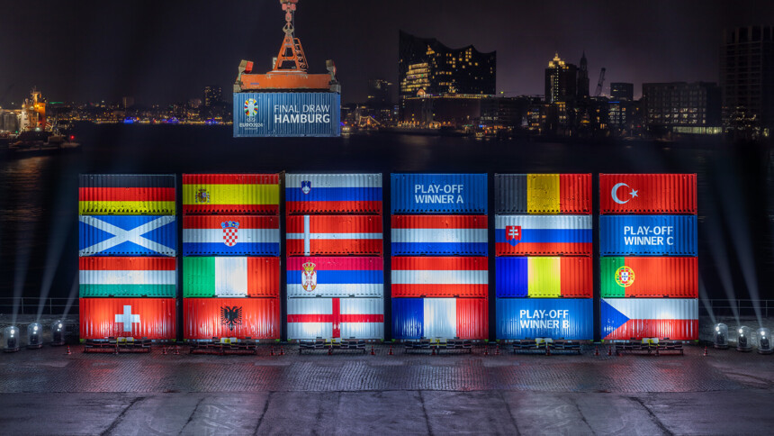 Night shot of the port of Hamburg with illuminated shipping containers showing the flags of the countries taking part in UEFA EURO 2024. A crane lifts a container labelled 'Final Draw Hamburg'. The Elbphilharmonie concert hall and the Hamburg skyline can be seen in the background.