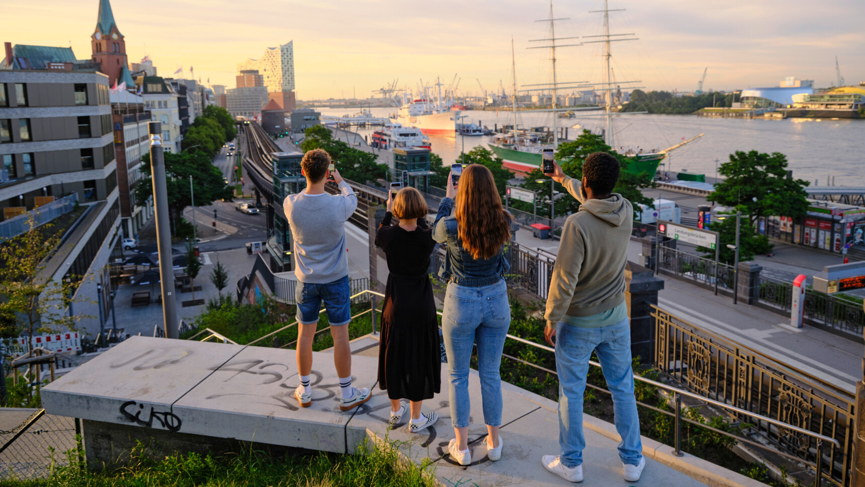 Vier Personen fotografieren mit Smartphones den Hamburger Hafen bei Sonnenuntergang.