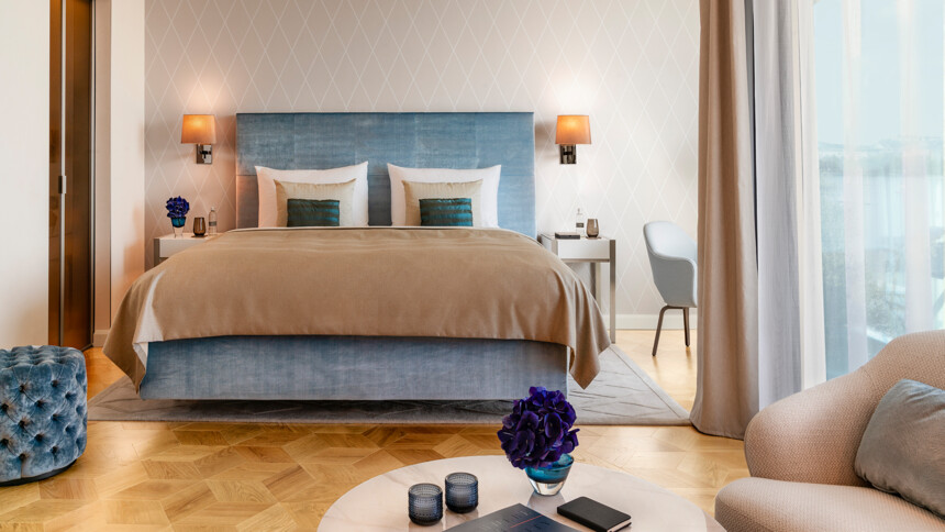 Modern eingerichtetes Schlafzimmer mit großem Bett, beigem Bettüberwurf, blauem Kopfteil und Tageslicht durch Fenster.