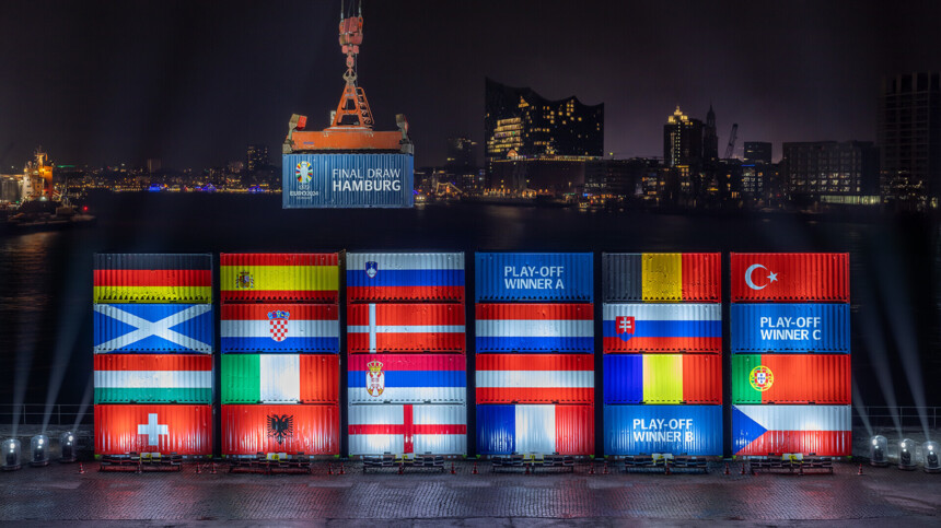 Night shot of the port of Hamburg with illuminated shipping containers showing the flags of the countries taking part in UEFA EURO 2024. A crane lifts a container labelled 'Final Draw Hamburg'. The Elbphilharmonie concert hall and the Hamburg skyline can be seen in the background.