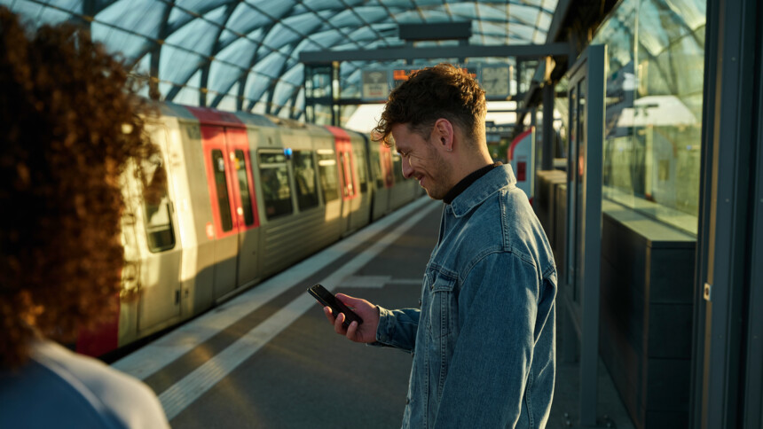 Ein Mann in Jeansjacke schaut lächelnd auf sein Smartphone an einem Bahnhof mit einfahrendem Zug.