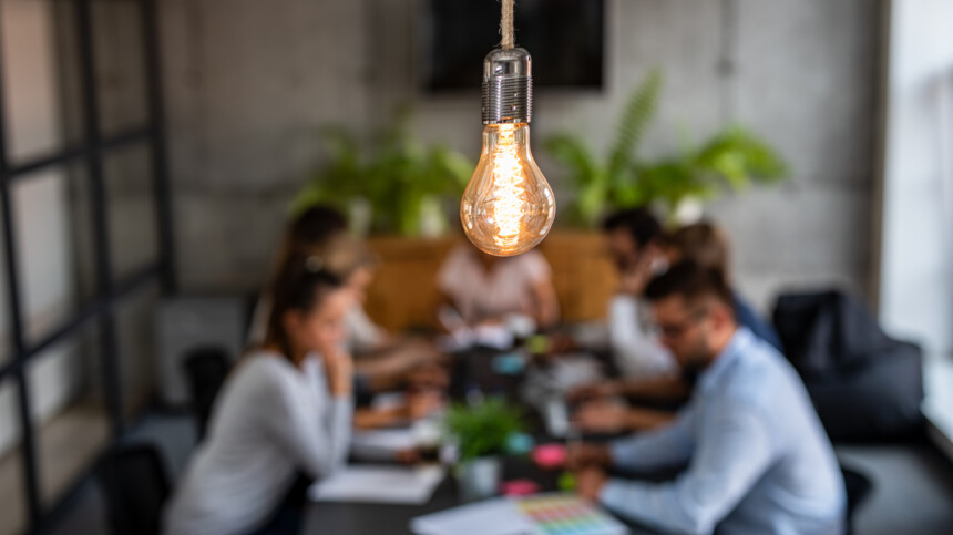 Leuchtende Glühbirne im Fokus, unscharfe Menschen bei Besprechung im Hintergrund.