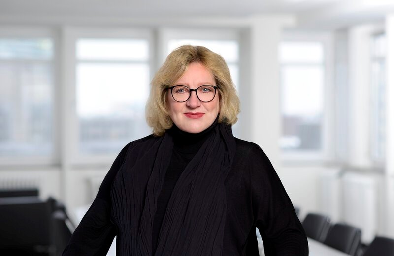 Porträt einer Frau mit blonden Haaren, Brille und schwarzer Kleidung in einem hellen Büro.