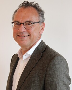 Porträt eines lächelnden Mannes mit Brille, grauem Sakko und weißem Hemd vor hellem Hintergrund.