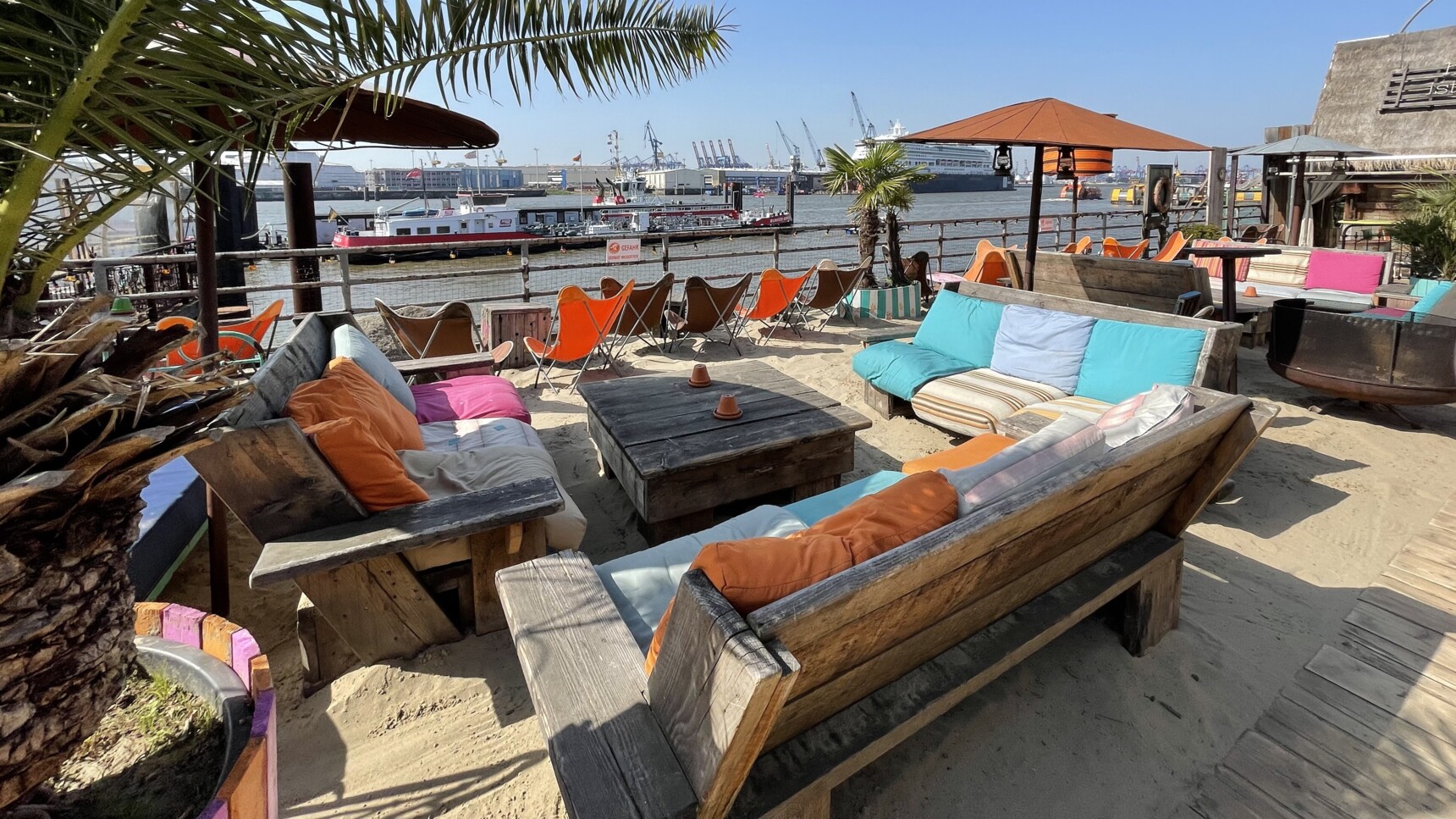 Strandbar mit bunten Sitzmöbeln, Palmen und Blick auf Hafen und Schiffe bei sonnigem Wetter.