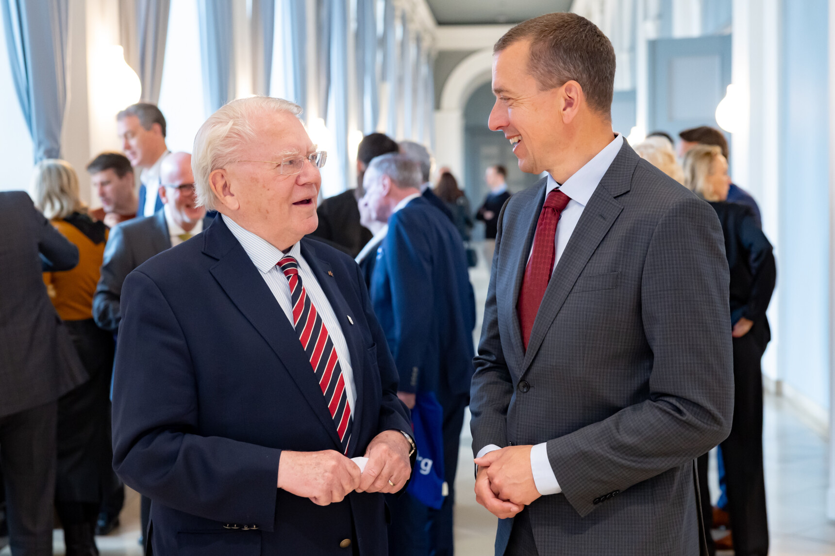 Zwei Männer im Anzug unterhalten sich freundlich in einem hellen, eleganten Flur mit mehreren Personen im Hintergrund.