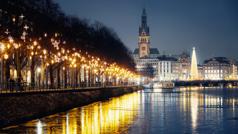 Weihnachtlich beleuchteter Alsteruferweg mit Rathaus und Weihnachtsmarkt in Hamburg bei Nacht.