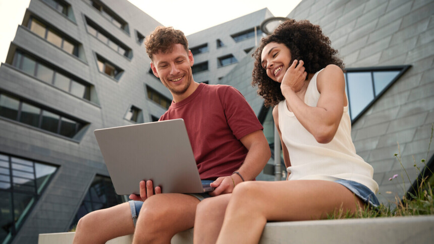 Zwei junge Menschen sitzen draußen vor modernem Gebäude und schauen gemeinsam auf Laptop.