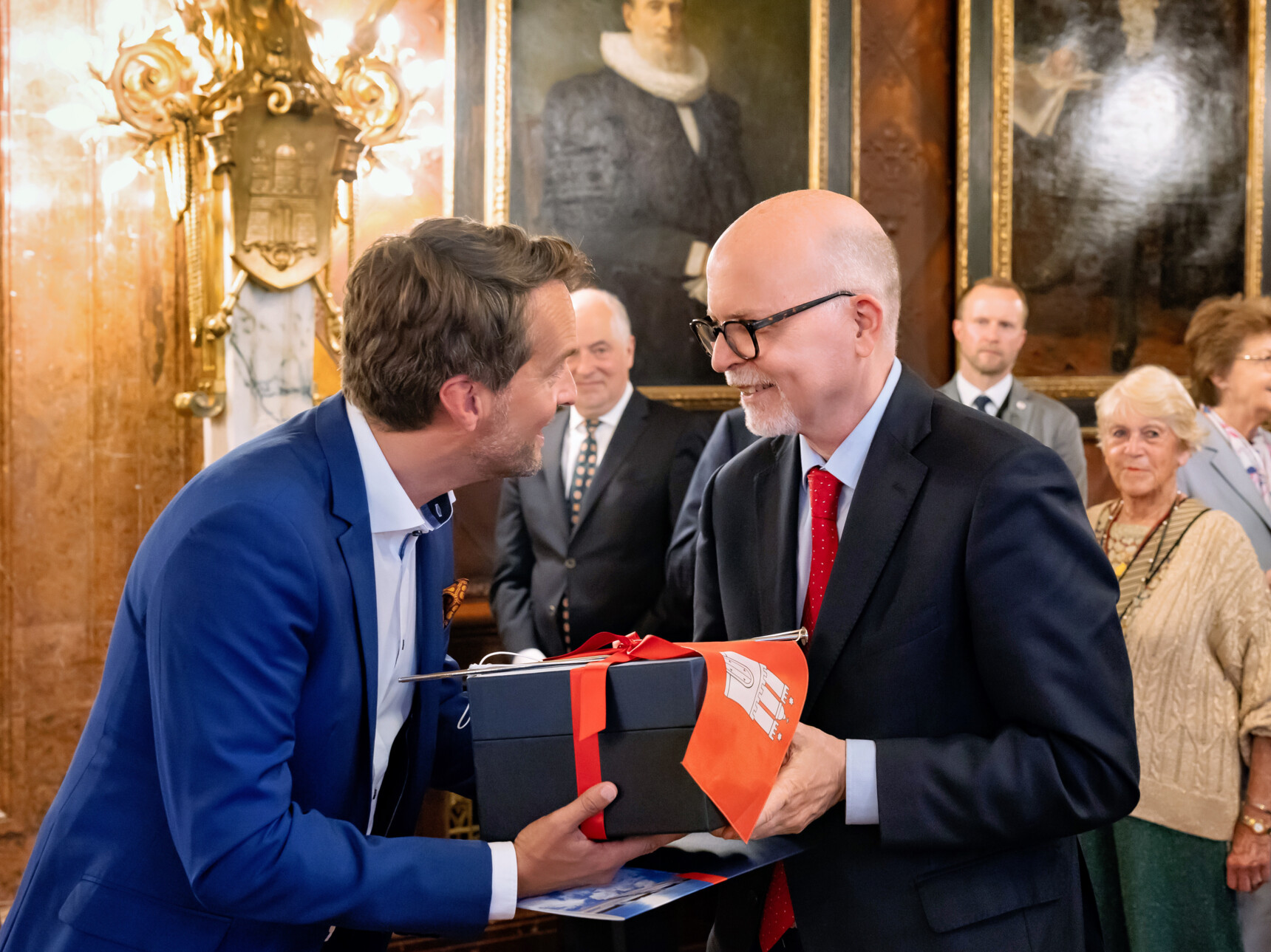 Zwei Männer im Anzug übergeben sich lächelnd ein Geschenk in einem festlichen, historischen Raum.